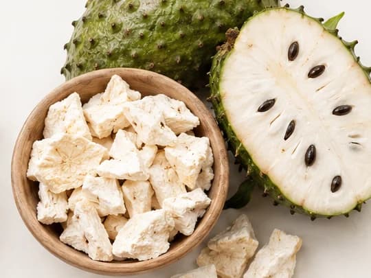 Dried soursop pieces in a wooden bowl beside a halved soursop